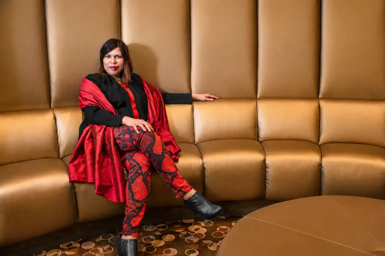 "Diane Rodriguez" wearing a bright red and black outfit sitting on a large cushioned bench looking into the camera.